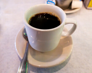A steaming cup of black coffee sits on a white saucer with a spoon, ready to be enjoyed. The deep, rich liquid has a slight foam on top, inviting a warm sip.
