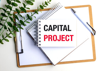 Notepad with capital project text on clipboard with pen and green plant