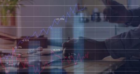 African American man penning in notebook at office desk with laptop, mug, plant displaying charts