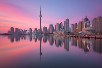 Fototapeta premium City skyline at dawn, reflected in calm water. Sunrise colors paint the buildings and sky. Iconic tower stands tall