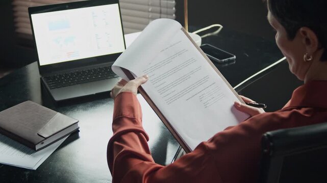 Cropped shot of mid-adult Caucasian woman signing document while sitting at desk in front of laptop with financial screen at office