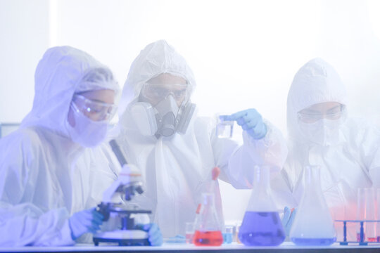 A diverse team of scientists collaborates in a high-tech biotech lab. A senior researcher mentors his colleague as she carefully pipettes a solution into a test tube for an experiment.
