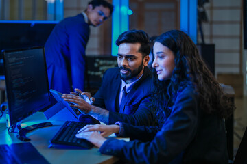 A diverse team of AI developers in a late-night coding session. A senior engineer uses a tablet to guide a programmer as they collaborate on building a complex neural network.