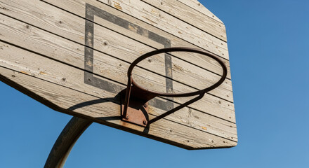 Weathered outdoor wooden basketball hoop with metal rim mounted against clear blue sky, representing urban sport nostalgia and recreational activity, dry sunny day