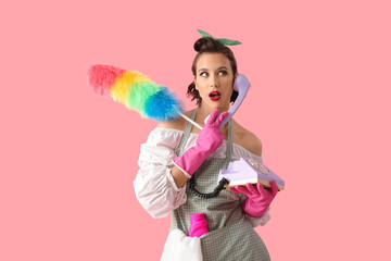 Beautiful young shocked pin-up housewife with pp-duster and detergent talking by phone on pink background