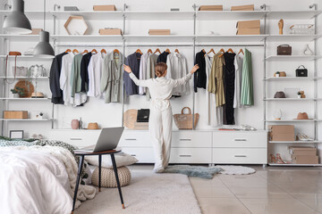 Young woman choosing clothes from wardrobe in light bedroom with cozy bed and laptop on bedside table, back view