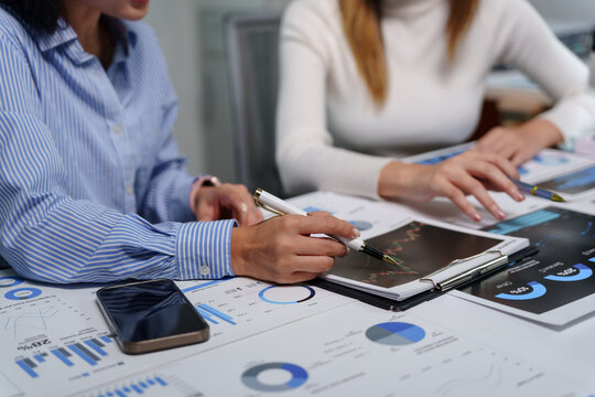 Businesswomen working together, analyzing financial data on charts and graphs, using pen and clipboard, in a modern office environment