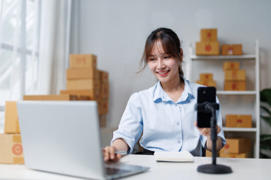 Young asian entrepreneur managing her online business using laptop and smartphone for live streaming, surrounded by cardboard boxes ready for shipment - Powered by Adobe
