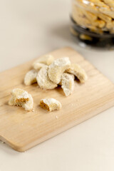 A pile of crescent-shaped putri salju cookies dusted with powdered sugar on a wooden board, with kastengel cookies in a plastic jar in the blurred background.