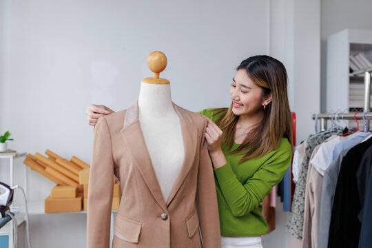 Smiling Asian fashion designer adjusting a blazer on a mannequin in her vibrant workshop, surrounded by creative garments and tools