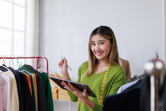 Asian female fashion designer using digital tablet and stylus pen managing online orders in her clothing store workshop