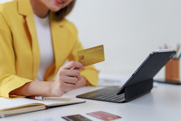 Businesswoman holding a gold credit card while using a tablet for online shopping, surrounded by a modern office setup at her desk