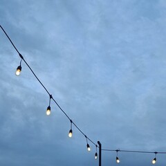 A string of hanging light bulbs glowing under a cloudy evening sky.