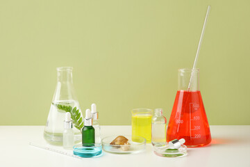 Laboratory glassware of colorful samples with Achatina snail and cosmetic products on white table near green wall