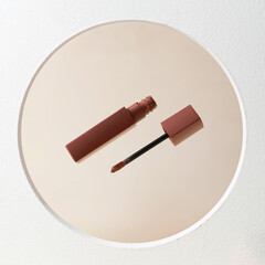A reddish-brown liquid lipstick with the applicator wand removed from the tube, positioned diagonally over a round mirror on a light beige background, creating a minimal and modern beauty composition.