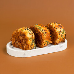 Three slices of savory bread roll topped with shredded meat floss, black sesame seeds, and green onion, arranged neatly on a white oval tray against a warm brown background.