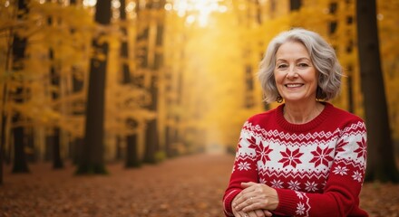 The smiling older woman in a red warm sweater poses in the autumn forest of gold leaves; a warm, hospitable portrait that transfers wisdom, family and seasonal comfort