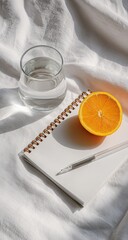 Sunlit still life of an orange half, a glass of water, a blank notepad, and a pen resting on a white fabric