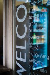 Transparent welcome sign sticker in various languages on the glass door of a food stall selling cold drinks with a refrigerator seen on the background