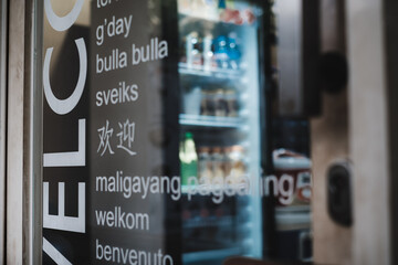 Multilingual welcome sign decal  on the transparent glass door of a tourist spot kiosk selling cold beverages with a refrigerator seen on the background