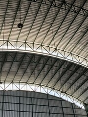 Close-up view of a large industrial metal roof with curved steel beams and structural trusses, featuring hanging lights in a spacious warehouse or factory interior.