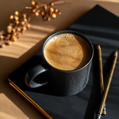 A dark mug of hot coffee sits on a black notebook with pencils and dried berries, evoking a cozy morning atmosphere