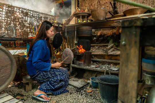 A Thai woman in traditional attire fans a charcoal stove in a rustic kitchen, capturing the warmth and charm of local culture.