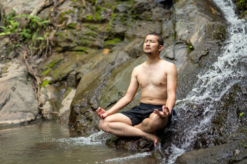 A shirtless man meditates in a lotus position beside a flowing waterfall, embracing peace, mindfulness, and natural healing in a serene outdoor setting.