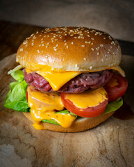 
A hamburger with a sesame seed bun, patty, melted cheese, lettuce, and sliced tomatoes sits on a wooden board. The background is dark.