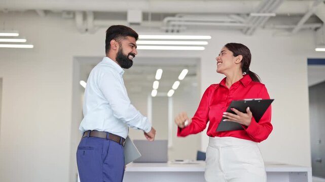 Successful Indian businessman and cheerful businesswoman, dressed professionally, celebrate exciting achievement with high-five in modern office, showing happiness and collaboration.