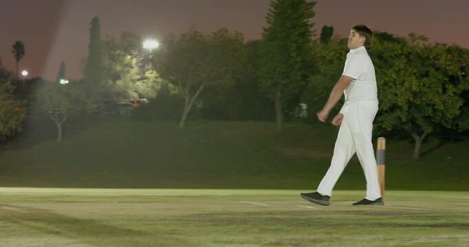 Delivering ball male bowler watching batsman leave crease resetting run-up on grass pitch in lights