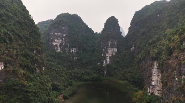 Towering limestone karsts aligned along the Red River Delta&rsquo;s edge encircle a serene river valley in Trang An, Ninh Binh, Vietnam, captured by ascending drone shot.