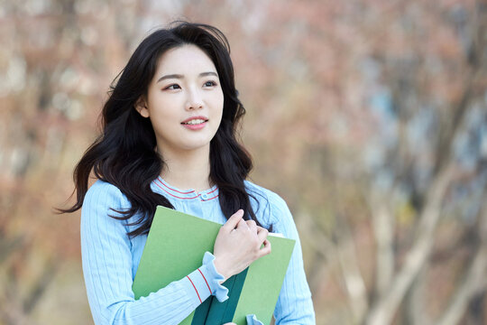 Young Asian female student holding books outdoors in springtime park
