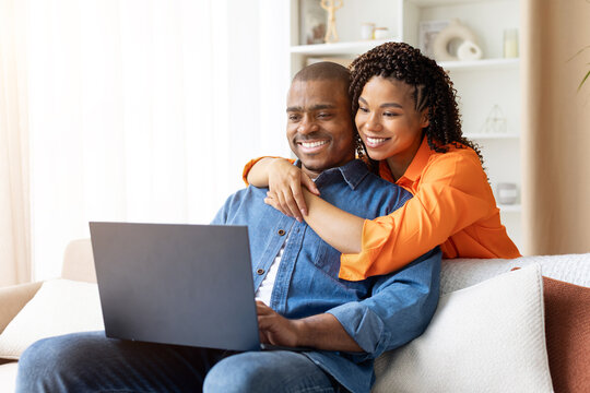 Happy young African American couple sitting on couch at home, smiling while using laptop together, woman in orange shirt hugging man from behind, copy space
