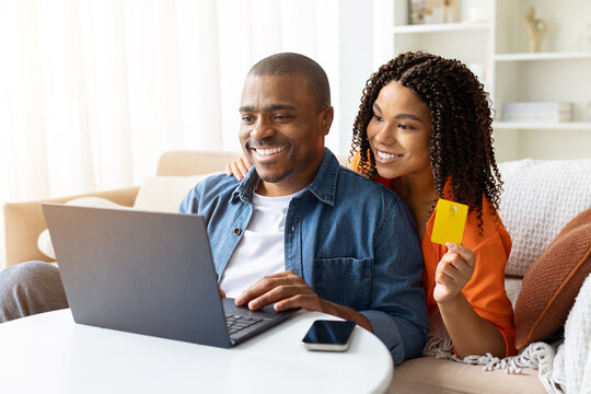 Happy young African American couple using laptop and credit card at home, cheerful black spouses smiling while shopping online together in living room interior