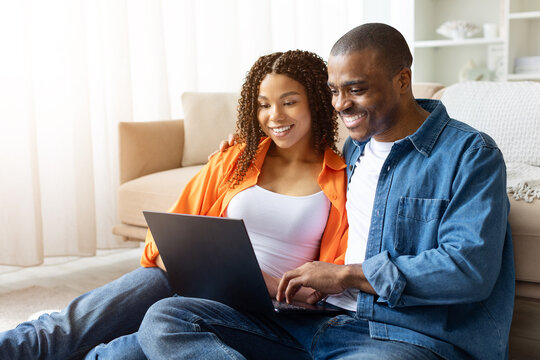 Smiling young African American couple sitting on floor with laptop at home, browsing together and enjoying time in casual relaxed setting, copy space