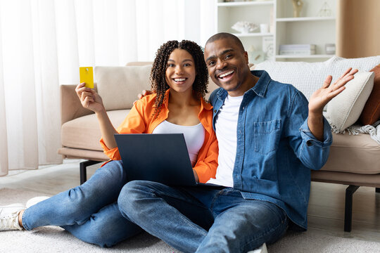 Excited young African American couple sitting on floor at home, holding credit card and laptop, smiling and celebrating online success together, closeup - Powered by Adobe
