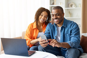 Smiling African American couple using smartphone together at home, sitting at table with laptop, looking at screen and enjoying moment.