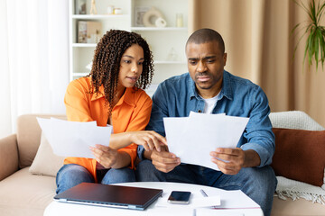 Serious young African American couple reviewing documents together at home, planning budget or checking mortgage papers, sitting at table with laptop and coffee cups, focused expressions