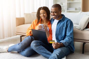 Joyful young African American couple sitting on floor at home with laptop, woman holding mug, man holding credit card while shopping online, copy space
