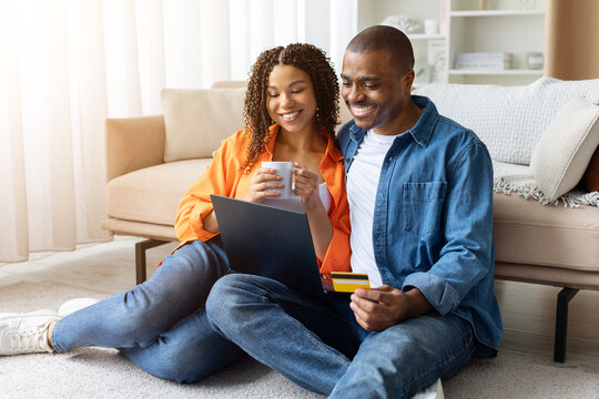 Joyful young African American couple sitting on floor at home with laptop, woman holding mug, man holding credit card while shopping online, copy space