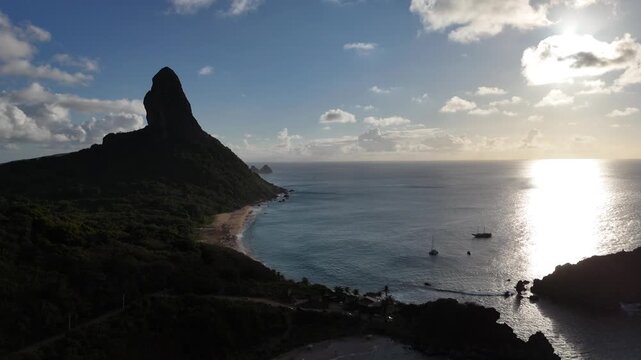Vista a&eacute;rea do Morro do Pico em Fernando de Noronha iluminado pela luz alaranjada do p&ocirc;r do sol, com o mar azul ao fundo. Uma cena espetacular que simboliza o encanto natural da ilha