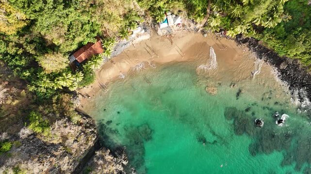 Aerial drone view of Praia do Cachorro, Fernando de Noronha, Brazil, highlighting turquoise waters, sandy beach, cliffs, and lush tropical scenery.
