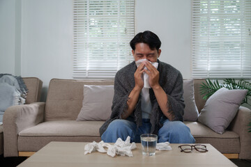 Sick asian man blowing nose while sitting on sofa at home