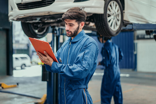 Mechanic in blue uniform reviewing service checklist on clipboard with colleague working in background, showing professionalism, attention to detail, and quality automotive service.