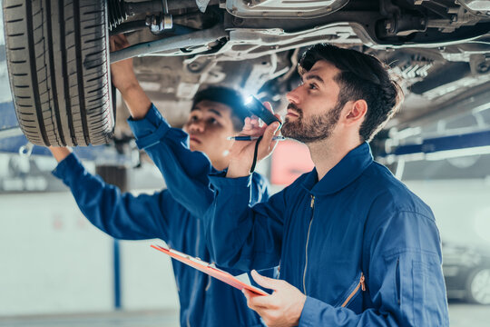 Two mechanics inspecting suspension and tire with flashlight and clipboard on lift, representing teamwork, maintenance, and repair expertise in automotive service. - Powered by Adobe