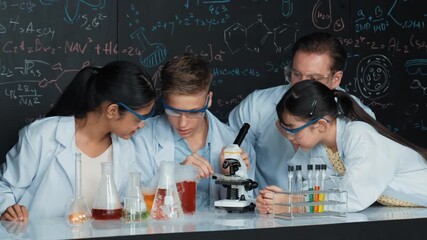 Multicultural student looking under microscope while cute girl inspect sample. Teacher looking after high school children doing experiment at blackboard with science theory in STEM class. Edification