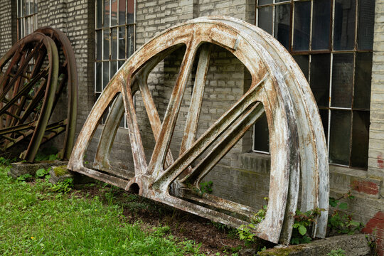 Large corroded steel pulley wheels from historic pithead machinery resting near a brick wall at an abandoned mining facility in Europe