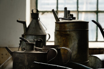 Old metal oil cans and jugs on a workbench in an industrial workshop, illuminated by window light, showing rust and patina