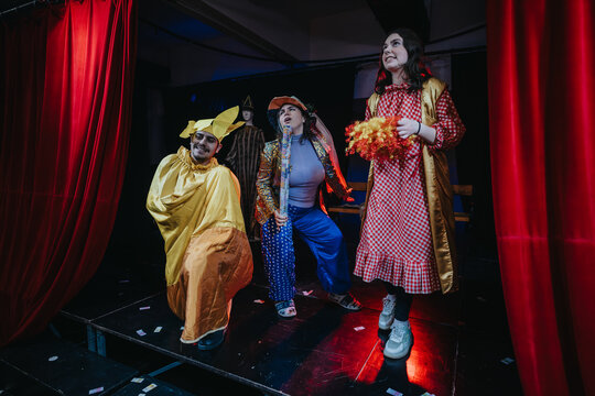 The image depicts three actors enthusiastically performing in vibrant costumes on a theatrical stage surrounded by red curtains, showcasing an exciting and colorful performance.